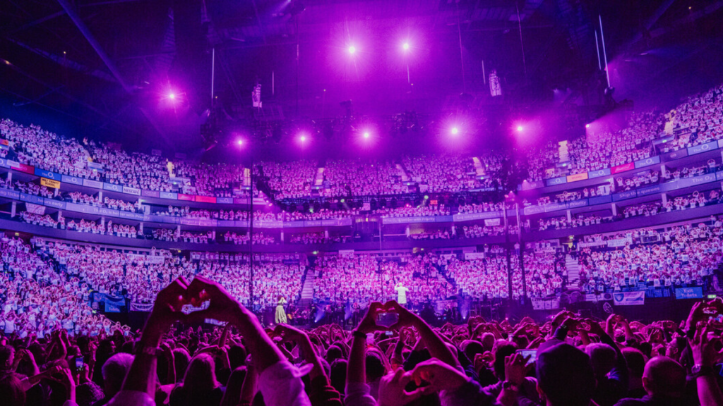 A photo of the O2 stage with audience with their arms raised.